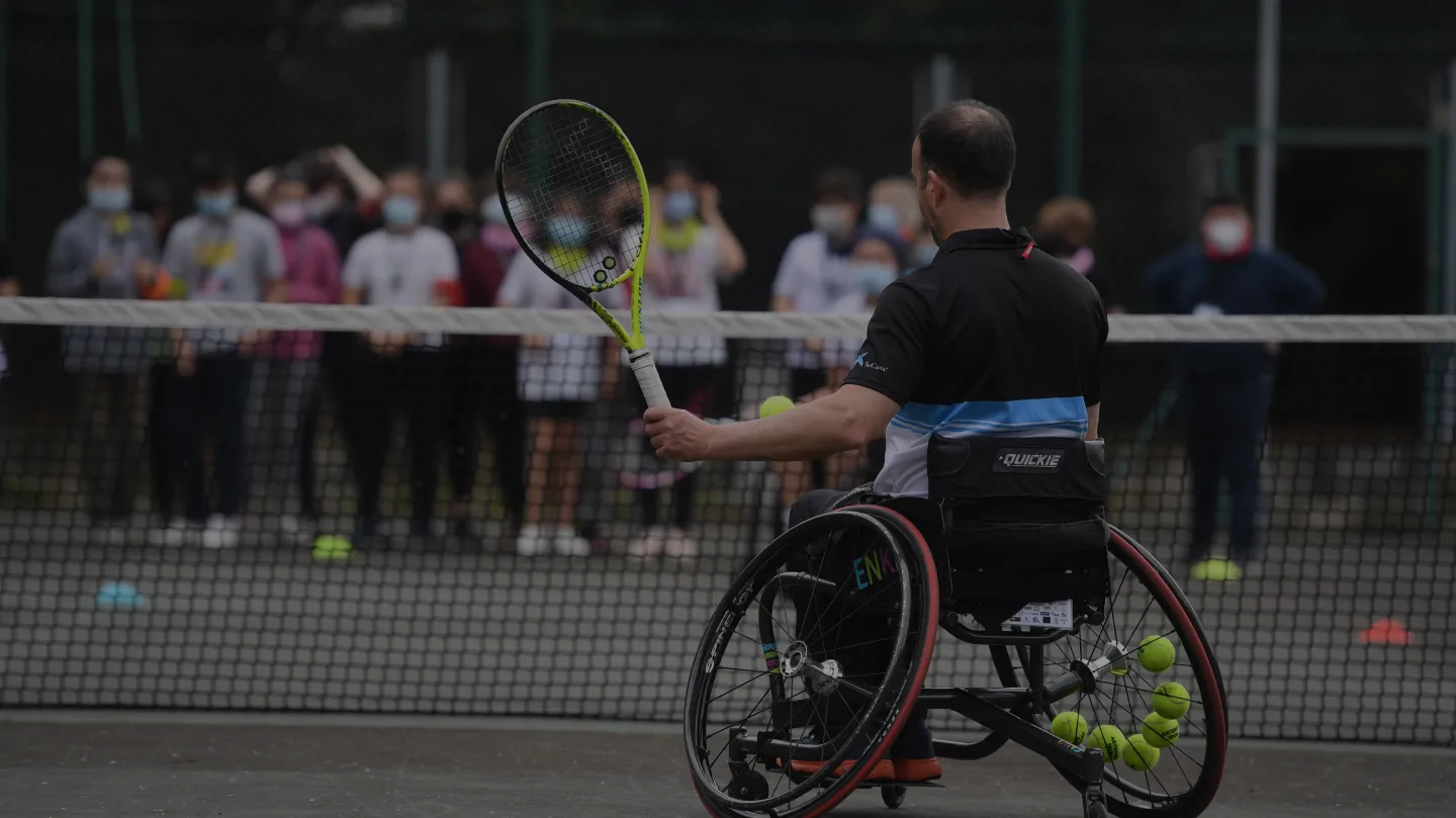 Persoa en cadeira de rodas xogando ao tenis mentres un grupo observa a actividade.